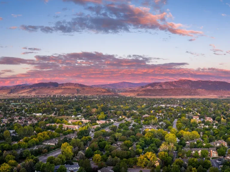 Suburban neighborhood with dense trees under a colorful sunset sky and distant mountains.