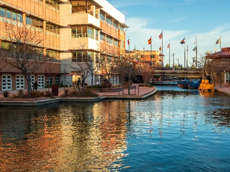 Urban canal with boats, flags, and modern buildings reflecting in the water at sunset.