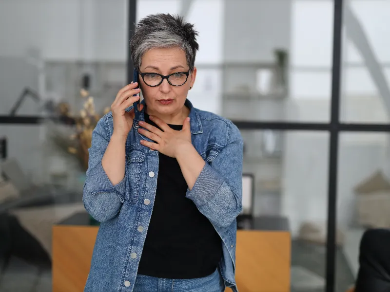 Middle-aged woman with short gray hair and glasses holds phone to ear and touches her throat, looking concerned.