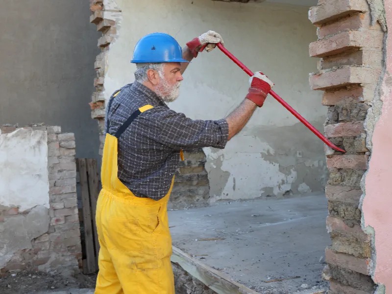 Construction worker in a blue helmet and yellow overalls using a crowbar to demolish a brick wall.