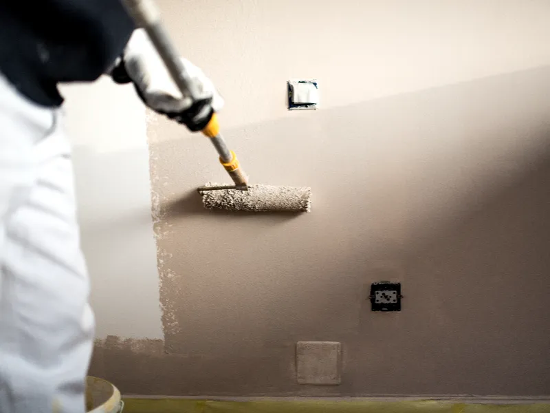 Person painting a beige wall with a roller brush.